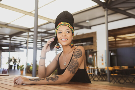 Beautiful tattooed African American woman wearing an african turban sitting at the table talking on her smartphone and smiling confidently. Copy space for text.