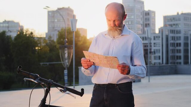 Grandfather male tourist of retirement age senior man city traveler standing on street outdoors with map and bike active elderly person searching way road destination navigation route looking around