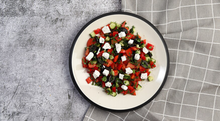 Greek salad with cucumbers, tomatoes, onions, olives, feta cheese and herbs on a round plate on a dark gray background. Top view, flat lay