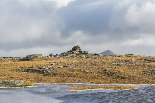 A small stony mountain sumit cairn with majestic mountain summit the background and small pond in the foreground under a stormy grey sky