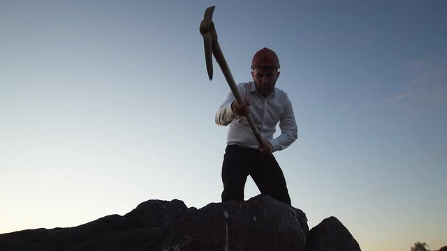 Caucasian man with a beard stands and hits the stones with a pickaxe, sparks and pieces of stone fly off against the background of outdoor sky. Mining concept goal achievement