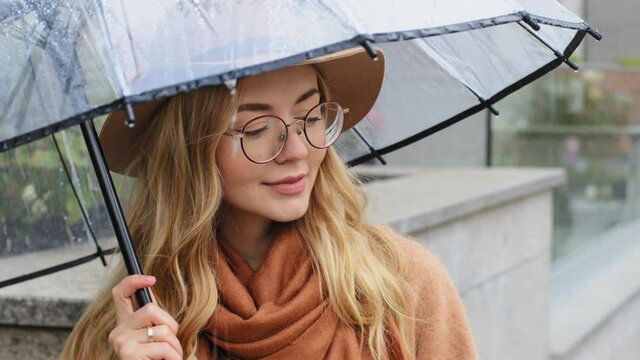 Portrait attractive girl standing with umbrella in city in rainy weather pensive young woman enjoying rain in autumn outside beautiful caucasian female smiling posing with hat and glasses on street