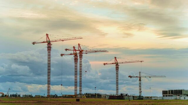Huge tower cranes operate with heavy building materials on construction site against distant city under cloudy sky from morning till night timelapse