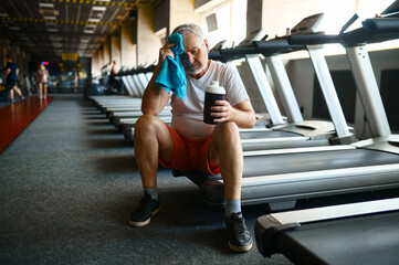 Fototapeta na wymiar Tired old man with towel drinks water in gym