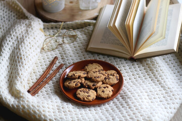 Plate of chocolate chip cookies, cinnamon sticks, soft blanket, open book and lit candles. Hygge at home. Selective focus.