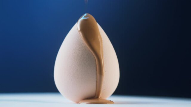 Foundation tone dripping on beauty blender sponge on blue background. Brown cream texture close-up. Makeup and beauty products concept, cosmetics, liquid bronzer or concealer.