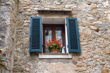 Fototapeta na wymiar A small window in a stone wall with flowers in a pot and dark aqua blue shutters in the medieval village of Dolceacqua, Italy.