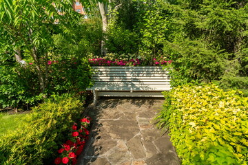 A resting bench stands in a secluded corner among flowers, trees and trimmed shrubs in a park on a sunny day in summer.