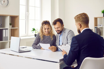 Happy clients sitting at desk in real estate agent's office and reading papers that he gave them. Young married couple who are planning to buy new house reading contract terms and conditions together