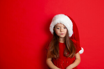 Merry Christmas and happy holidays. Portrait of a sad girl in a Santa cap on a red background. Kids, children. A place for text.