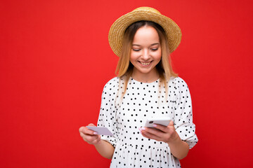 Photo of cheerful pretty blonde woman in summer outfit using mobile phone and holding credit card making payment online isolated over red background. Copy space