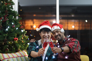 Adorable smiling young LGBT couple sharing special moment together on Christmas holiday, Asian gay male lover sitting on sofa in living room, drinking champagne to celebrate night before Christmas.