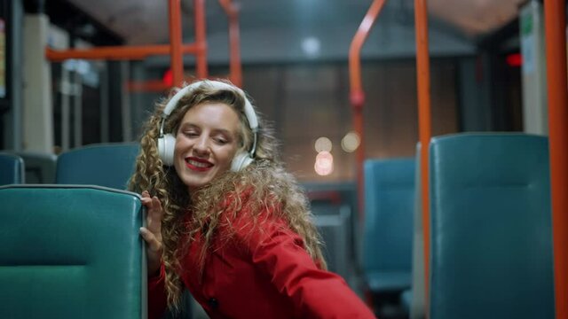 A smiling curly woman in headphones is listening music and funny dancing in bus. Girl passenger at modern tram. Public transport and happiness or fun concept. Night time. City lights background.