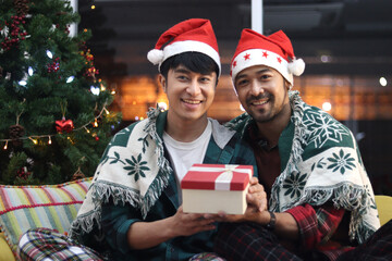 Adorable smiling LGBT couple holding present box, two men having special warm moment together on Christmas holiday, Asian gay male lover sitting on sofa in living room to celebrate Christmas night.