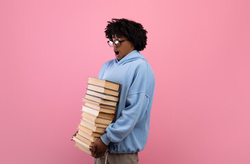 Overwhelmed black teenage student holding big stack of books, having lots of homework on pink...