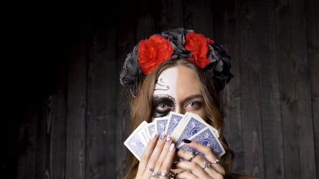 Lady model with skull makeup in red and black wreath choker and in lacy gloves shows gaming cards against wooden studio wall closeup