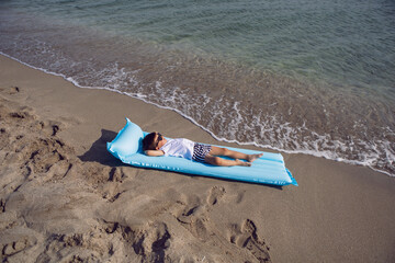 boy child in a white T-shirt and striped swimming trunks and sunglasses is lying on a blue inflatable mattress on the beach the sailor