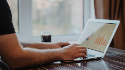 Close-up of a man who is typing on a laptop and working with a finance charts on a messy wood office table.