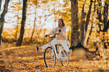 Young pretty woman riding vintage white bicycle in autumn park. Lady having fun on orange nature fall background.