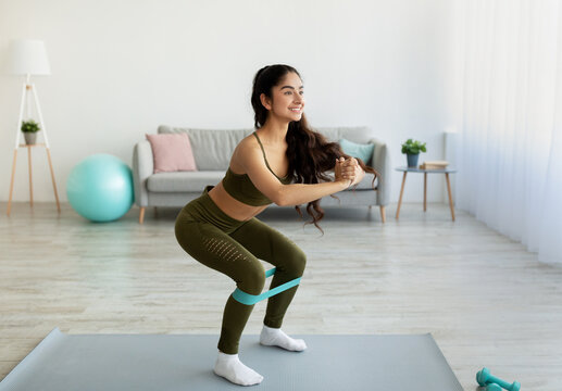 Fit young Indian woman doing squats with elastic band during home workout, copy space. Domestic sports concept
