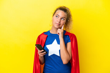 Super Hero woman isolated on yellow background using mobile phone and thinking