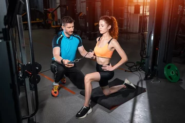 Tuinposter young woman exercising single arm row kneeling with personal trainer man in gym © goami
