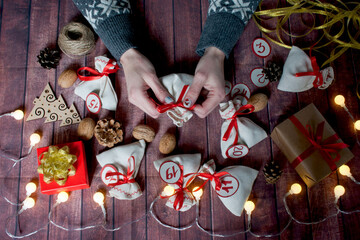 Christmas advent calendar. Woman holding bag with gift in her hands.
Bags with gifts, walnuts, cones, soutache, ribbon, boxes, garland, numbers, wooden Christmas tree are laid out on wooden surface