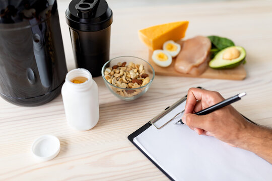 Closeup of young bodybuilder writing meal plan on clipboard with mockup, eating healthy foods, using protein shakes