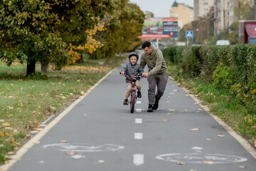 Father teaches his son to ride a bicycle on the bike path in the park. The father is holding a bicycle and the son is sitting on it