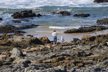 Mãe e filha bebé juntas na praia perto da água do mar - paradas a observar a água - família -...