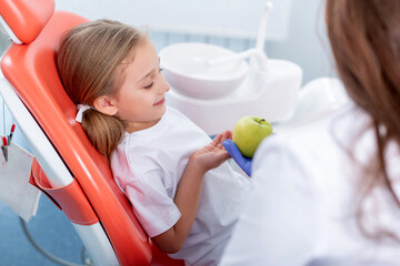 child girl in white clothes at reception at a pediatric dentist smiles and laughs. dentist gives the girl a big green apple. concept is children's health.