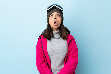 Mixed race skier girl with snowboarding glasses over isolated blue background looking up and with surprised expression.