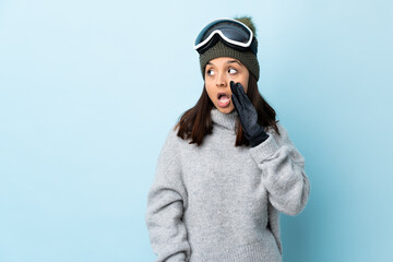 Mixed race skier girl with snowboarding glasses over isolated blue background whispering something with surprise gesture while looking to the side.