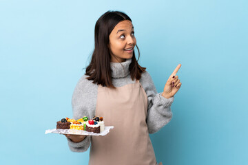 Pastry chef holding a big cake over isolated blue background pointing up a great idea.