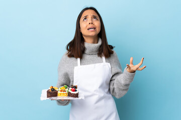 Pastry chef holding a big cake over isolated blue background stressed overwhelmed.