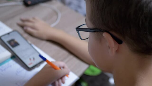 Schoolboy in Eyeglasses Writes with Pen, looking in Smart Phone. A left-handed child is sitting homework at the desk with books. Online a lesson with a gadget. Education concept. 4K. Close up.
