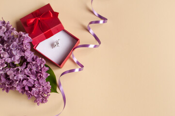 A white gold ring with a large gemstone in an open red gift box with a bow on a beige background next to a purple lilac branch with green leaves and a purple serpentine with a place for text