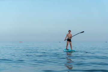 A boy of 11 years old swims on a SUP board in the sea after sunset.