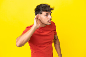 Young caucasian handsome man isolated on yellow background listening to something by putting hand on the ear