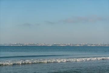 Sea waves running on a sandy shore on a clear sunny day. City in the background.