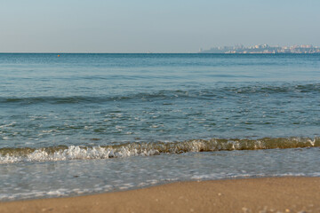 Sea waves running on a sandy shore on a clear sunny day. City in the background.