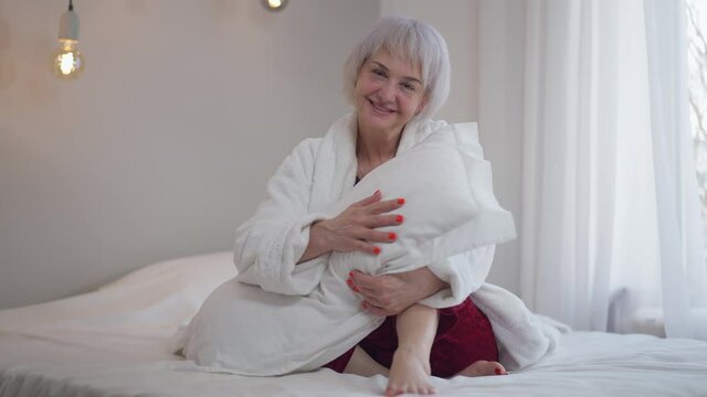 Happy smiling mature woman smelling fresh clean pillow looking at camera sitting on bed. Wide shot portrait of cheerful slim lady posing at home indoors. Hygiene and cleanliness concept