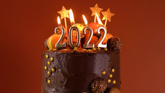 Happy New Year's Eve 2022 chocolate cake decorated with gold burning candles on aqainst dark wood table setting background. Close up cinemagraph loop static.