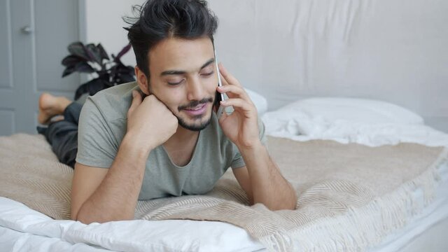 Young attractive Arab man is chatting on mobile phone in bed in apartment enjoying conversation. Sociable youth and communication concept.