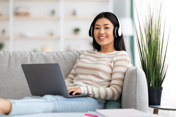 Naklejka premium Portrait Of Smiling Asian Lady Wearing Wireless Headphones Using Laptop At Home