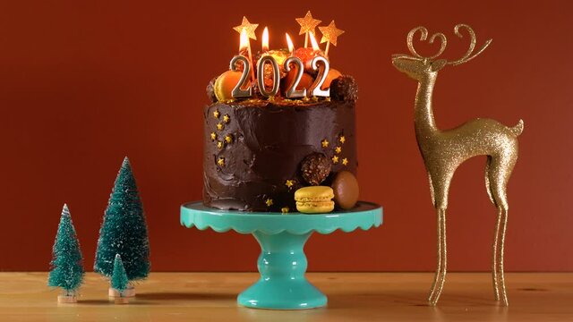 Happy New Year's Eve 2022 chocolate cake decorated with gold burning candles on aqainst dark wood table setting background. Cinemagraph loop static.