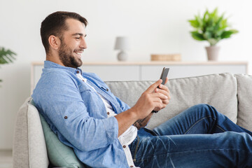 Cheerful adult european guy with beard sits on sofa chatting on phone in living room interior, side view