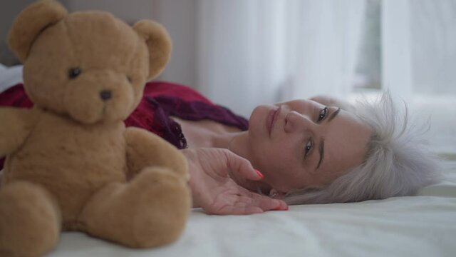 Side view happy woman lying on white bed smiling looking at camera with blurred teddy bear at front. Portrait of relaxed beautiful slim mature Caucasian lady posing in bedroom at home indoors