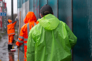 Workers in bright raincoats walk along the road along the fence. Rainy day, city views. Traffic. Three people, a view from the back.