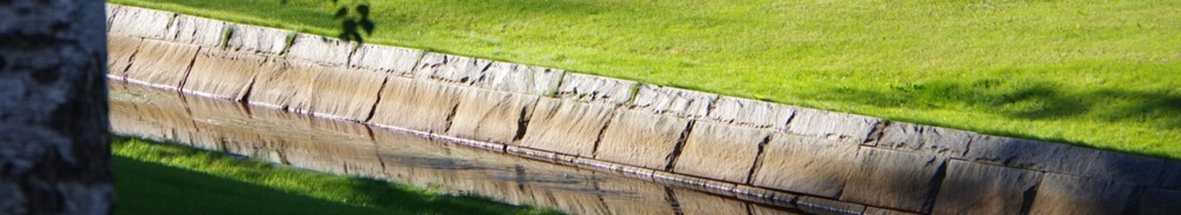 Fototapeta premium Narrow canal in public park with green grass meadow riverbank gutter banner